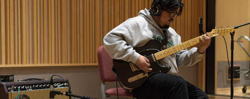 Male student wearing headphones playing a guitar in recording studio.